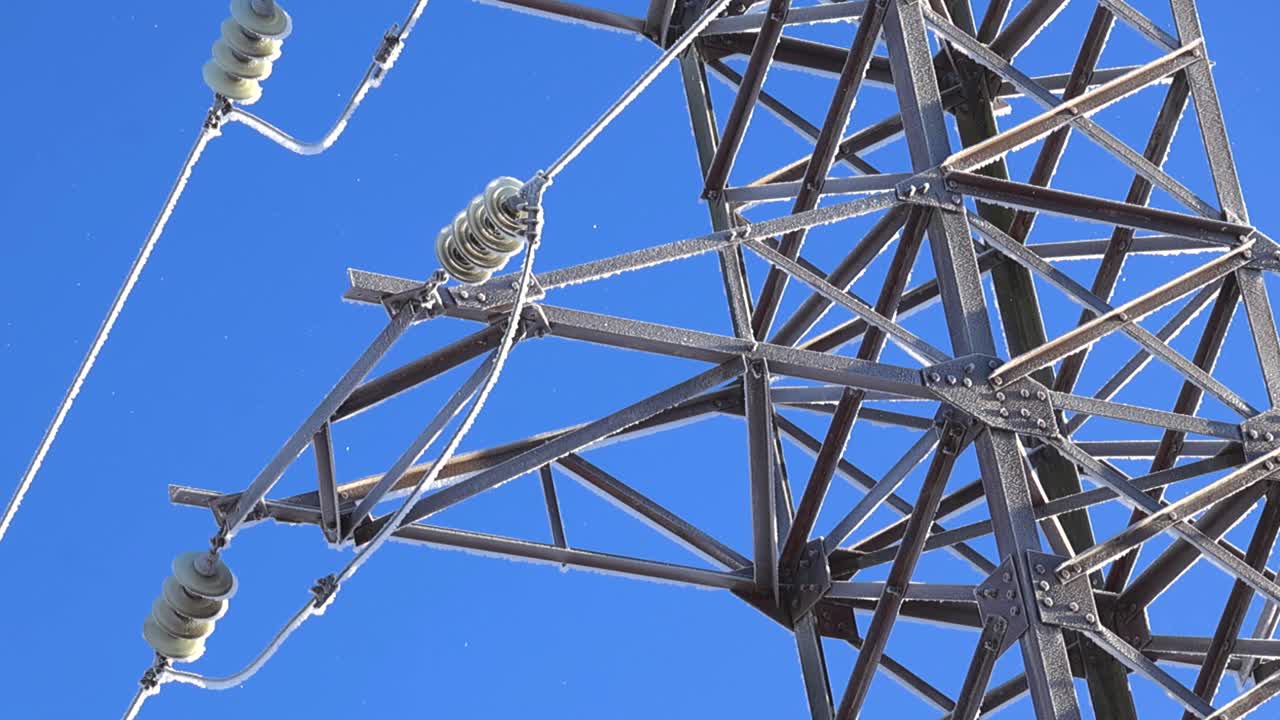 Torre de transmissão e cabos gelados cintilam sob o sol forte enquanto neve leve flutua pela imagem