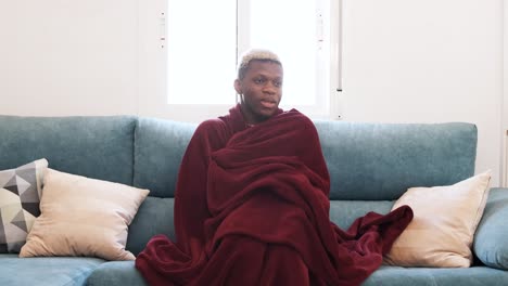 young man wrapped in a blanket on a sofa, appearing cold or unwell