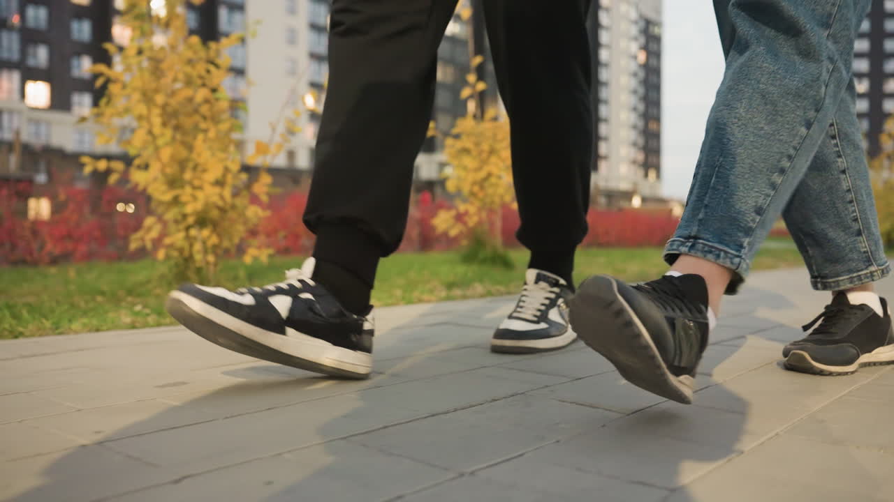 Leg view of people walking together on paved path, shoes in motion, with their shadows casting on ground, trees and modern buildings visible in background as they stroll through city streets