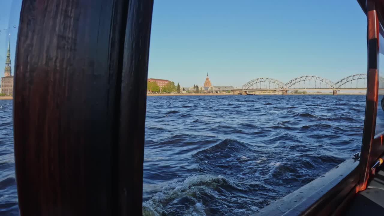 Boat-side view gliding along the Daugava River, framed by wooden rails, with Riga’s Old Town spires and the arched railway bridge appearing across lively blue waves