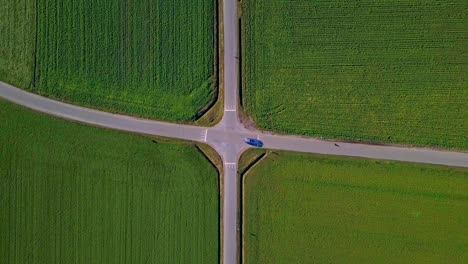 Vista-Aérea-Perfecta-De-Un-Cruce-Simétrico-Con-Campos-Verdes-Y-Un-Coche-Pasando