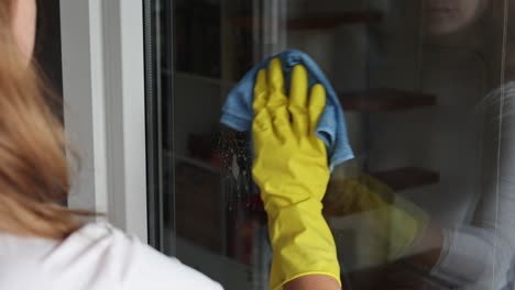 woman, housework and housekeeping concept - woman in gloves cleaning window with rag and cleanser spray at home