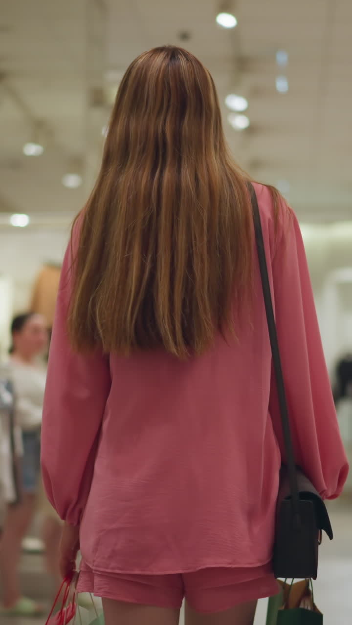 백인 여성이 두 개의 쇼핑 가방을 들고 선반과 선반에 끔하게 배열된 옷을 가진 옷 가게를 걸어가는 뒷면, 배경에서 지나가는 두 명의 여성의 약간 흐릿한 시각