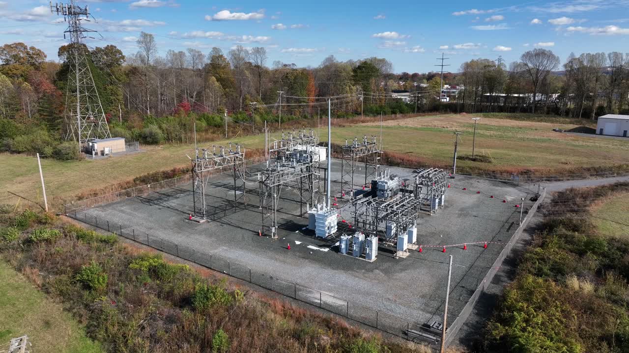 Aerial approaching shot of American electrical substation surrounded by open fields and autumn trees, showing metal structures, transformers and power lines under a clear blue sky