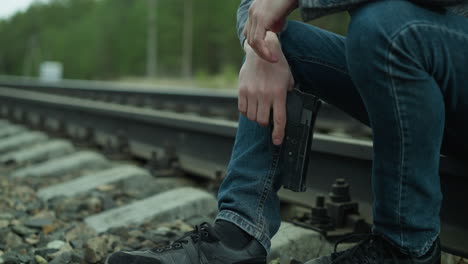 a close view of a man in a gray jacket, blue jeans, and canvas shoes, handling a handgun while sitting beside a railway track, the background shows a blurred view of a green forest