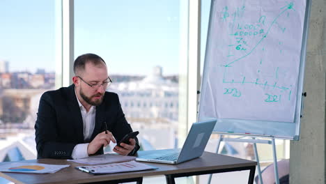 annoyed businessman sitting at desk with a phone. middle-aged entrepreneur in glasses using smartphone and talking to himself in office. business problems.