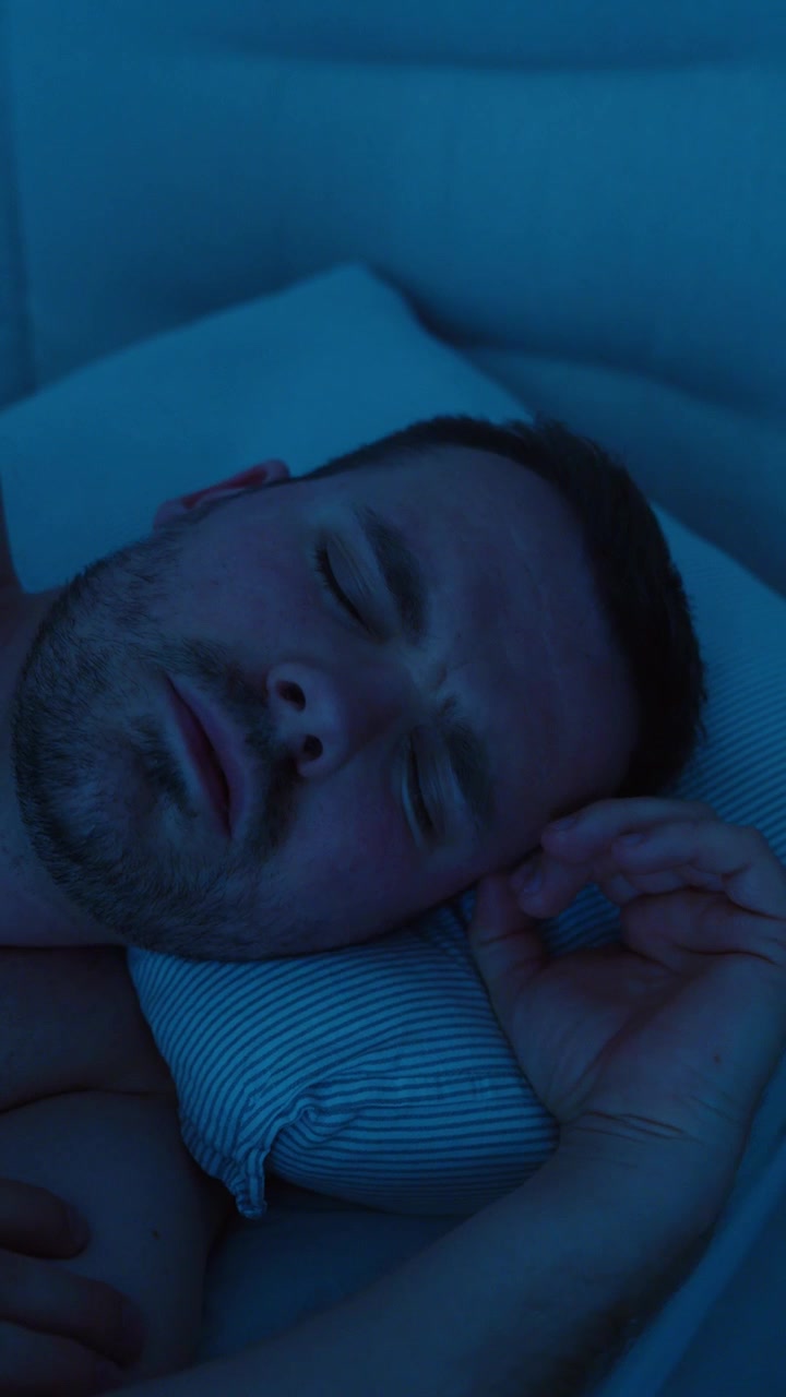 Uomo sdraiato a letto di notte che muove la testa mentre dorme irrequieto, suggerendo scarsa qualità del sonno, disagio, insonnia, stress o disturbi del sonno.