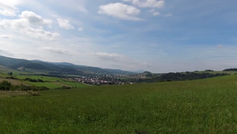 Langsamer-Schwenk-Von-Rechts-Nach-Links-über-Eine-Grüne-Wiese-Mit-Stadt-Und-Bergen-In-Der-Ferne-Mit-Blauem-Und-Bewölktem-Himmel