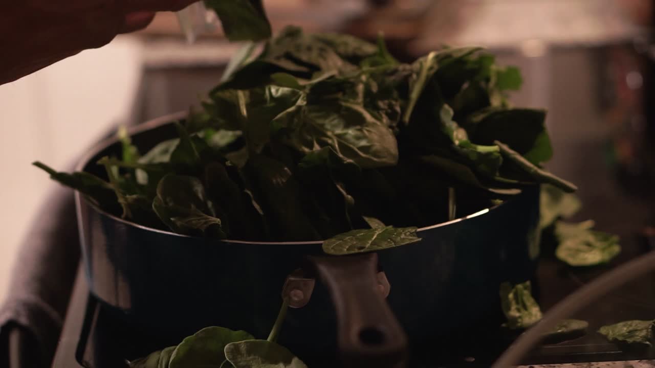 dropping and piling up spinach in deep blue pan on glass top stove with some spilling over