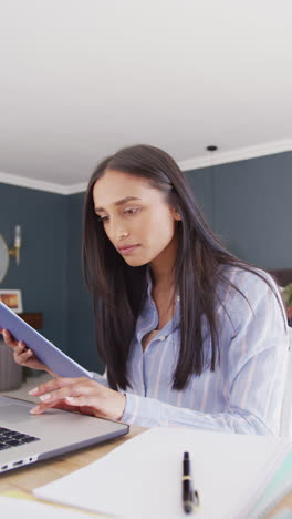 video vertical de una mujer biracial trabajando en el dormitorio usando una computadora portátil y leyendo notas