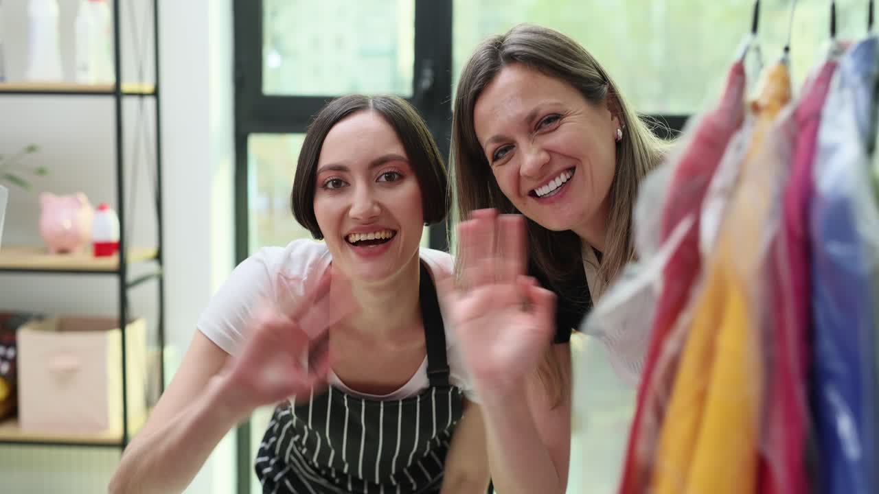 Mujeres sonrientes saludando desde detrás de un perchero de ropa en una tintorería.
