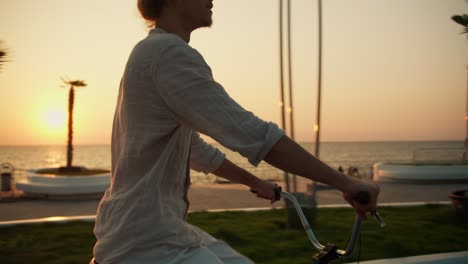 a happy young guy in a light shirt rides a bicycle along the beach where palm trees grow near the sea at sunrise in summer. a guy does morning exercises and drives along the beach in the summer