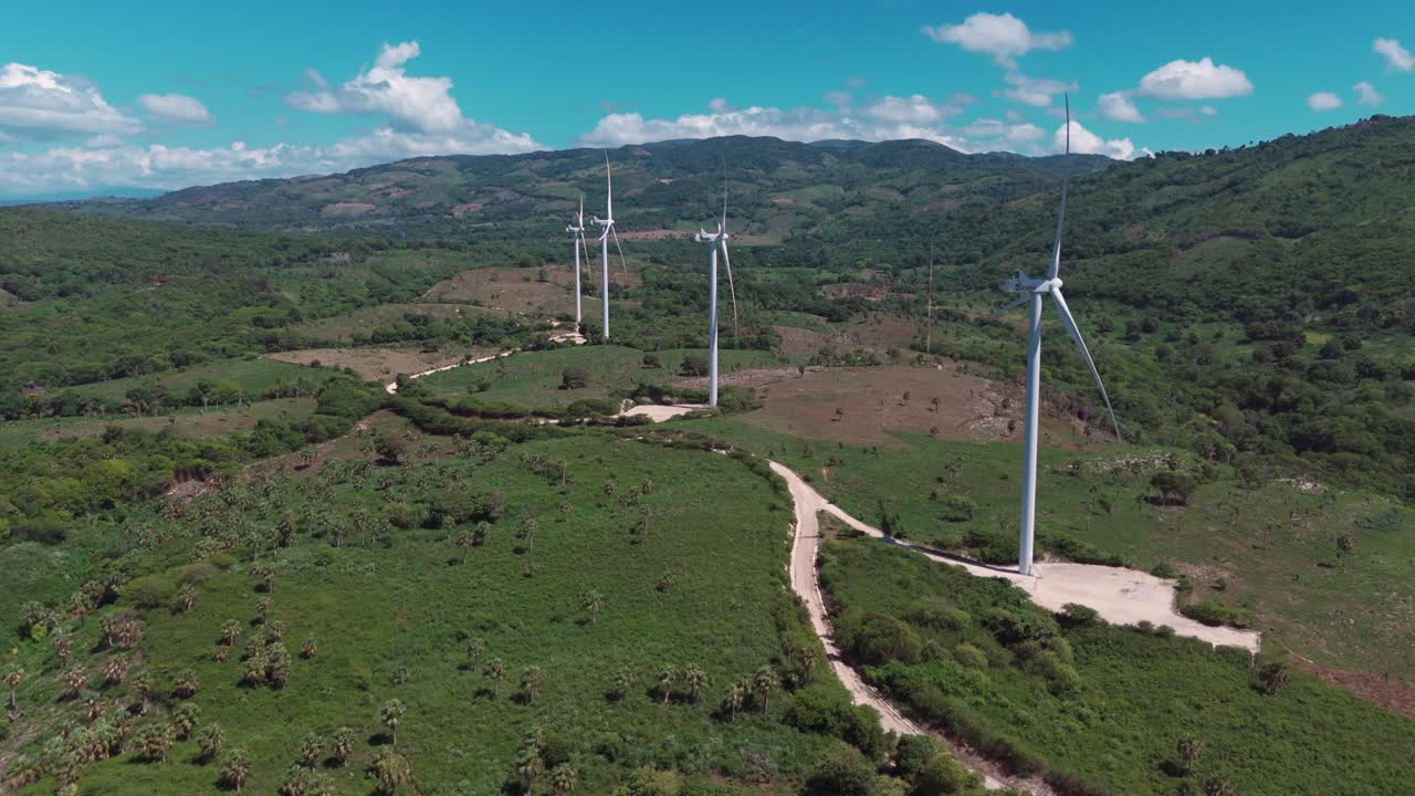 Aerial view of road in green hills of wind park with many wind turbines producing green energy. Sunny Summer day in Barahona, Dominican Republic. Wide shot