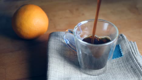 Cámara-Lenta-De-Café-Negro-Que-Se-Vierte-En-Una-Taza-De-Espresso-Italiana-Transparente-Sobre-Un-Trozo-De-Tela-Gris-Junto-A-Una-Naranja-En-El-Fondo