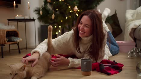 Happy-brunette-girl-in-a-white-sweater-lies-on-the-floor-on-the-Red-Reach-and-strokes-her-cream-colored-cat-in-a-cozy-room-decorated-in-Christmas-style