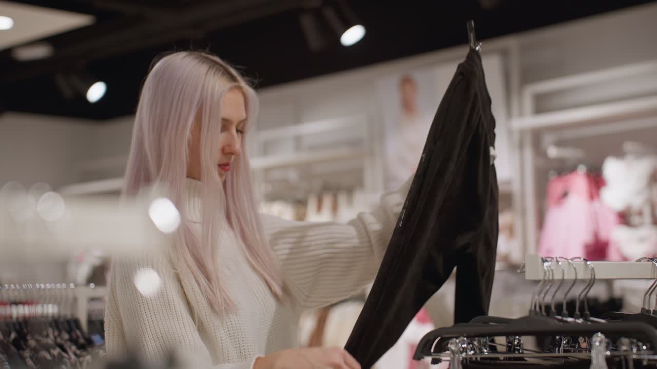 Passionate shopper in white knit sweater examines black jogger trousers on plastic hangers in bright boutique, feeling fabric texture and assessing fit before placing it back on rack with care