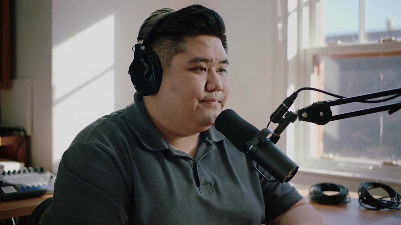 Người dẫn chương trình đang tựa người trả lời câu hỏi ngoài máy quay trong studio, mặc áo polo tối màu và đeo tai nghe, sử dụng micrô.