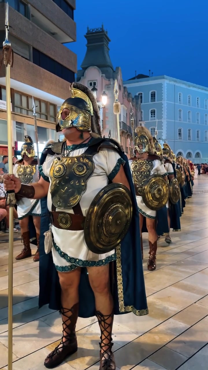 Alicante, Spagna, 8 agosto 2025: Uomini in armatura da soldato romano che marciano durante una parata serale a Cartagena. Gruppo di rievocatori con elmi e corazze in ottone che trasportano scudi e lance per strada.