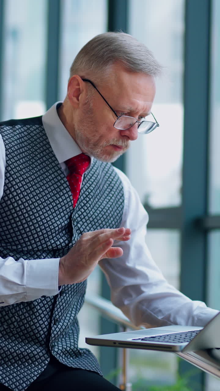 Nervous mature man working on a laptop. Serious senior businessman in eyeglasses standing near the window and looking at his laptop in his office. Vertical video