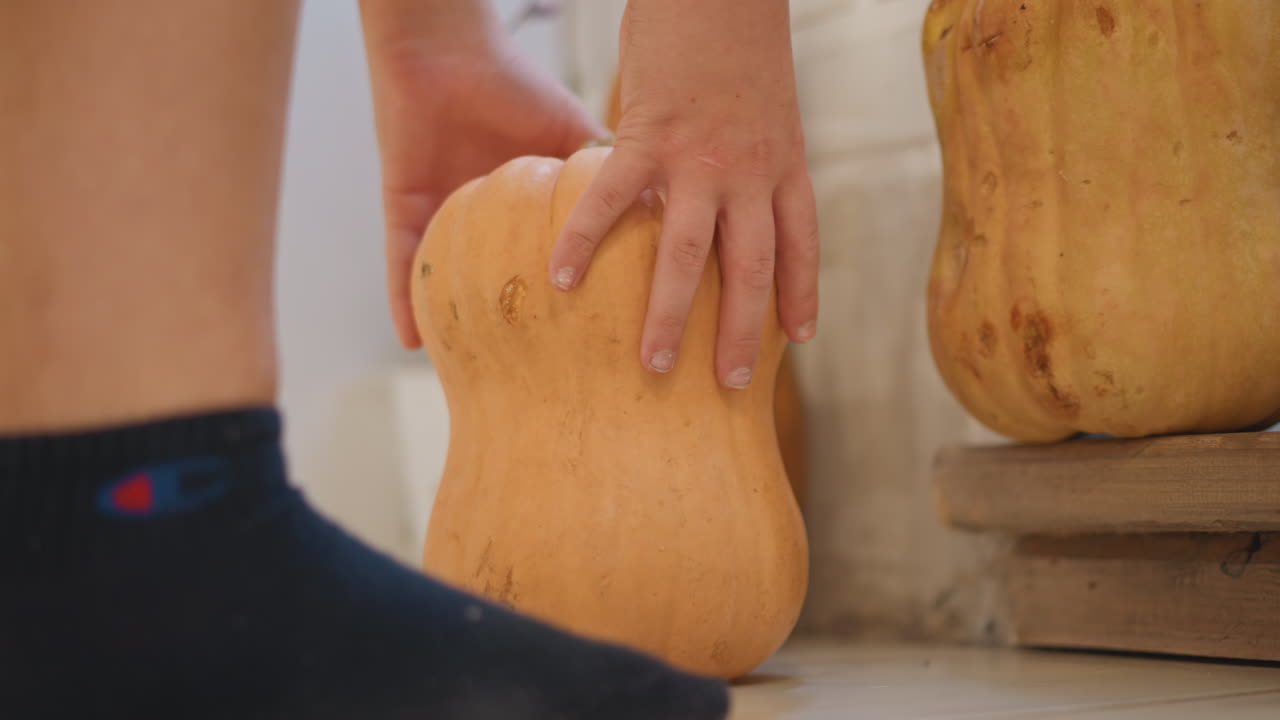 Bambino/a che si allunga verso una zucca su piastrelle, piccolo piede calzato che spinge la zucca mentre la mano afferra la buccia curva, scoperta giocosa in un angolo della cucina, luce calda suggerisce un momento di preparazione dei prodotti della raccolta, preparazione in famiglia per la cottura