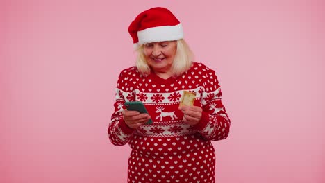 Ältere-Weihnachtsgroßmutter-Mit-Kreditkarte-Und-Smartphone,-Die-Geld-Beim-Online-Einkauf-überweist