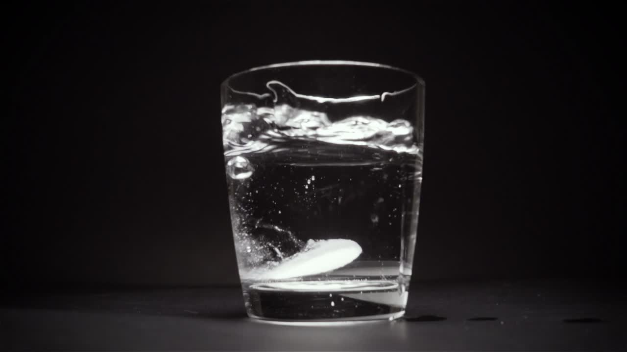 Una compressa cade sul fondo di un bicchiere d'acqua, sfondo nero, slow motion.