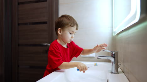 boy in red t-shirt washes his toothbrush in the sink. kid touches his tongue with toothbrush.