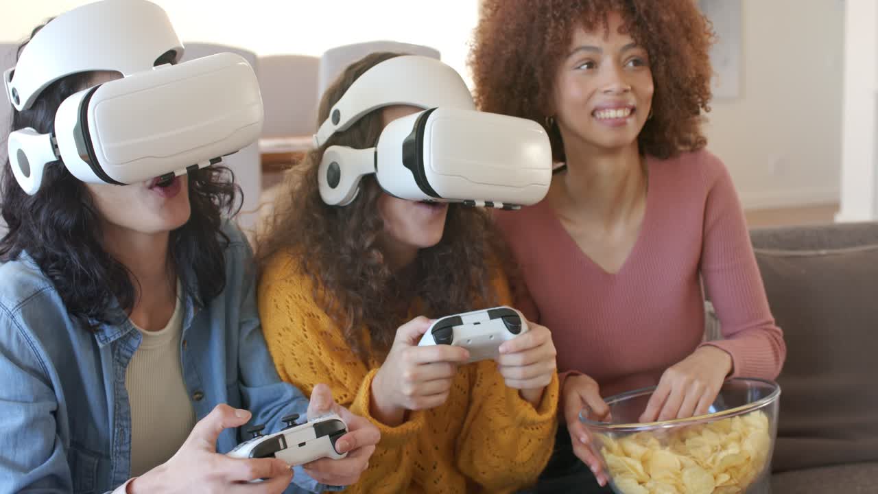 Diverse female friends donning VR headsets grabbing controllers on gray sofa gaming, snacking chips