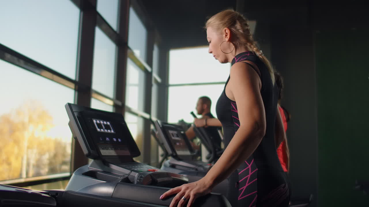 A woman walks up to the treadmill and turns it on to start training. Include a treadmill in the gym fitness room.