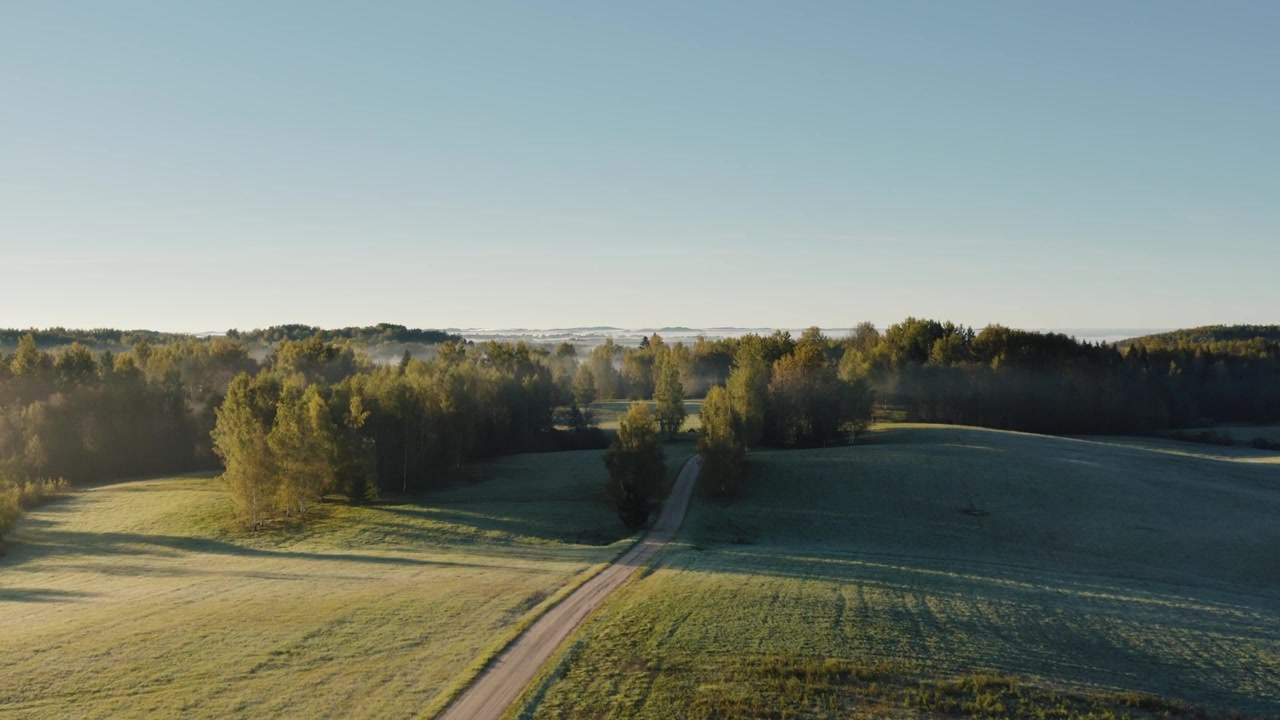 Drone shot over a countryside gravel road in early sunny morning with fog breaching in the landscape.