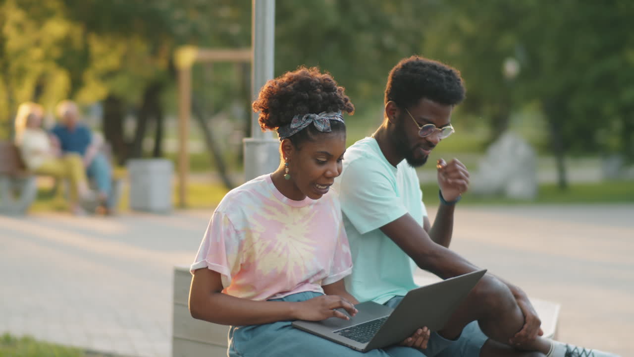 African American Woman Using Laptop and Talking with Friend on Bench in Park