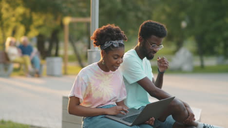 african american woman using laptop and talking with friend on bench in park