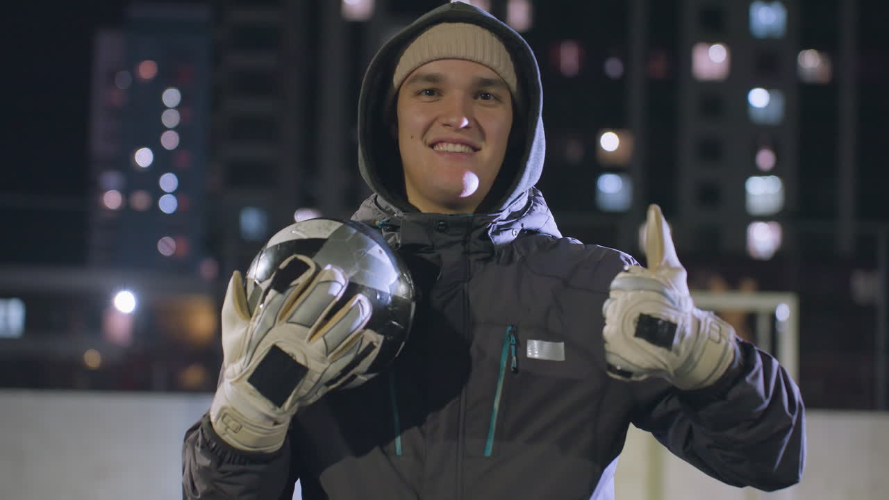 미소 짓고 엄지손가락을 올리며 축구공을 들고 장갑을 입은 젊은 축구 선수, 보케 조명과 함께 도시 야간 배경