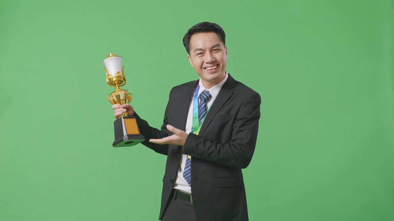 슈트와 타이를 입은 아시아 사업가, 손에 금메달을 들고, 손에 황금 트로피를 가리키며 미소 짓고, 스튜디오의 녹색 스크린 배경에서 첫 번째 우승자로 자랑스럽게 승리합니다.