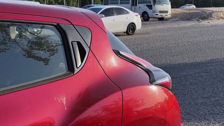 Primer plano de excrementos de pájaro sobre la pintura roja brillante de un coche aparcado. El vehículo está en un aparcamiento exterior soleado en Jabriya, Kuwait, con un sedán blanco y un autobús al fondo