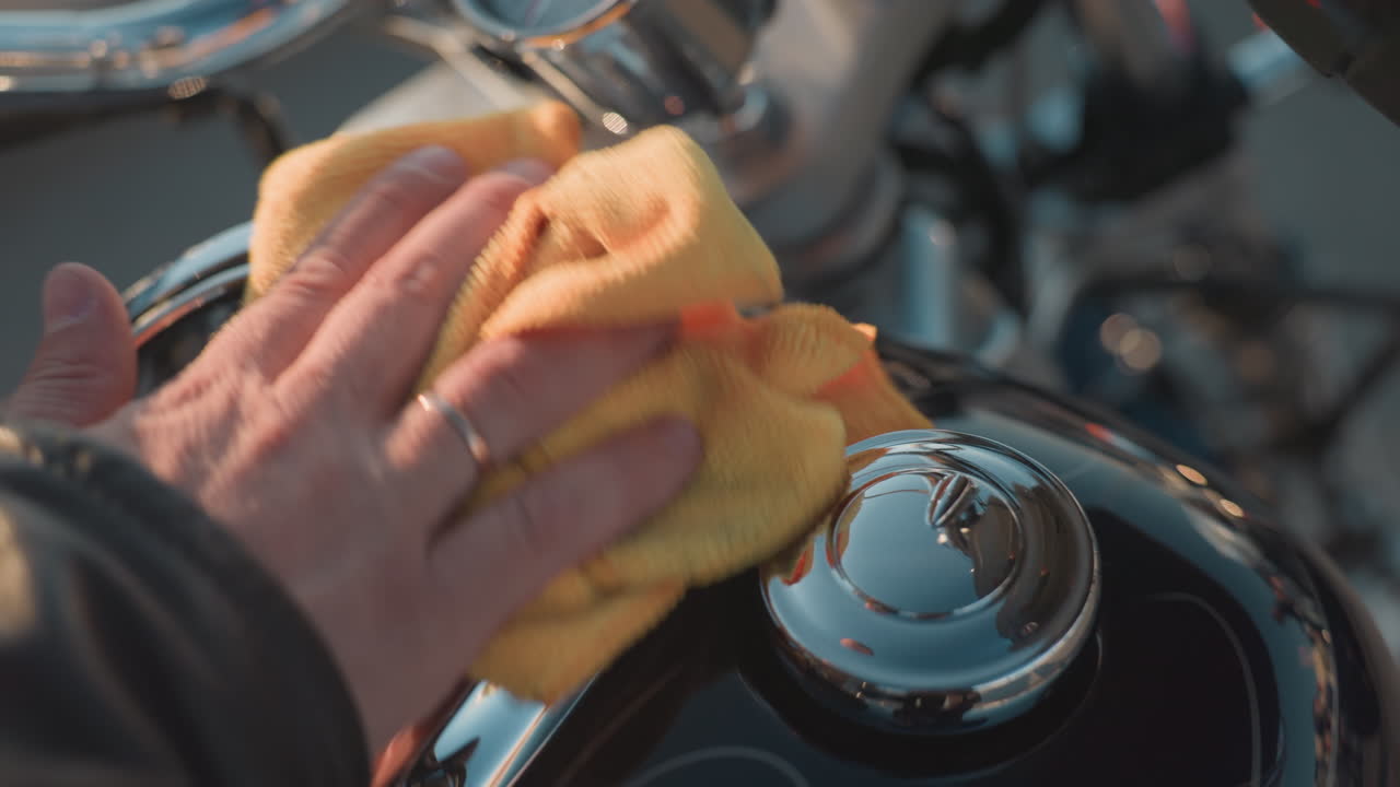 Close up of biker hand in leather jacket cleaning motorcycle with yellow cloth, focusing on careful maintenance of chrome and handle details under sunlight for polished