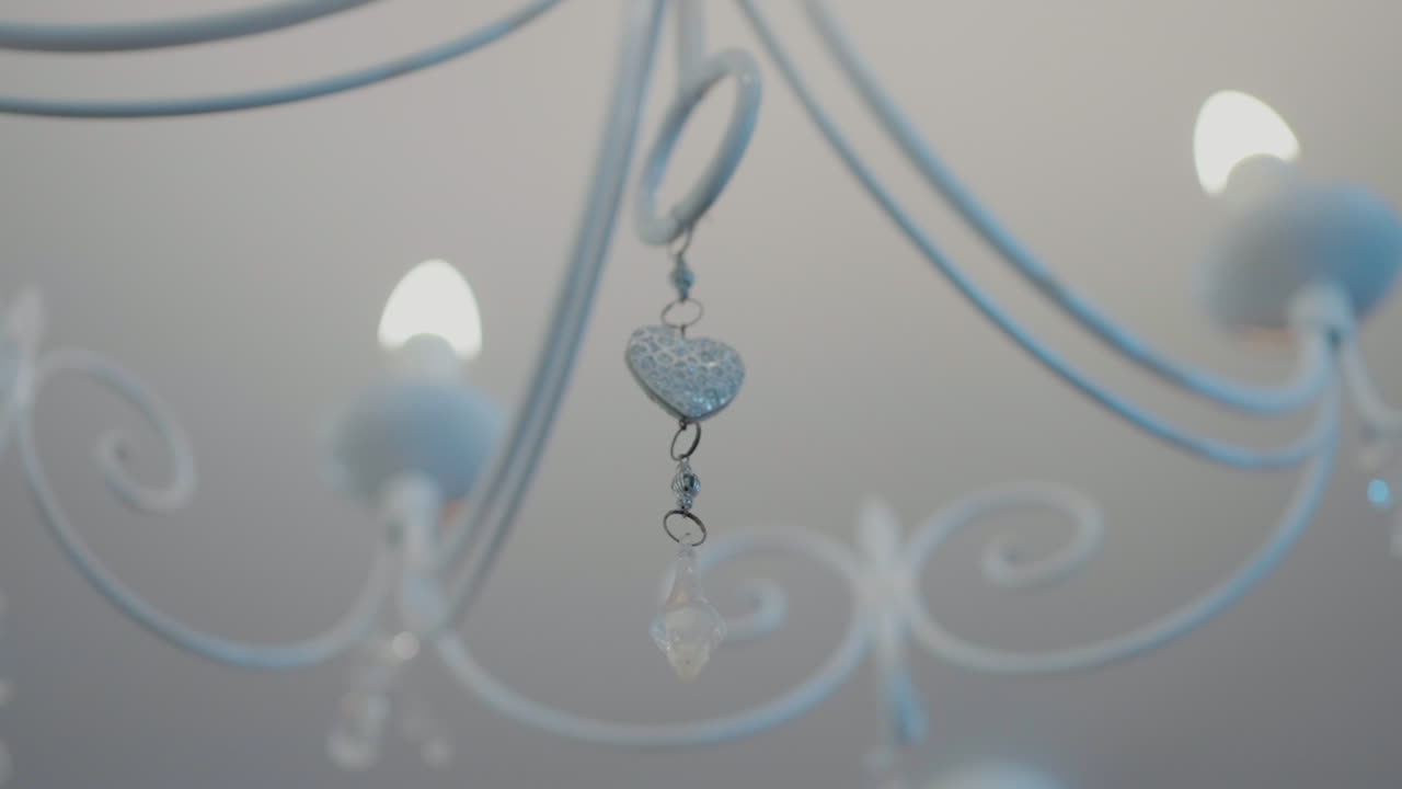 Primo piano di un lampadario bianco con delicati cristalli e ornamenti a forma di cuore, che emette una luce soffusa.