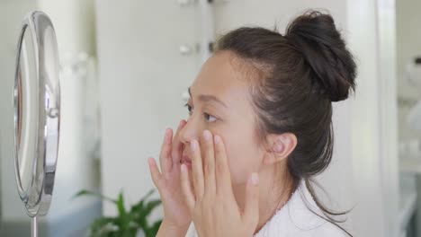 happy asian woman looking at mirror and touching face in bathroom, in slow motion