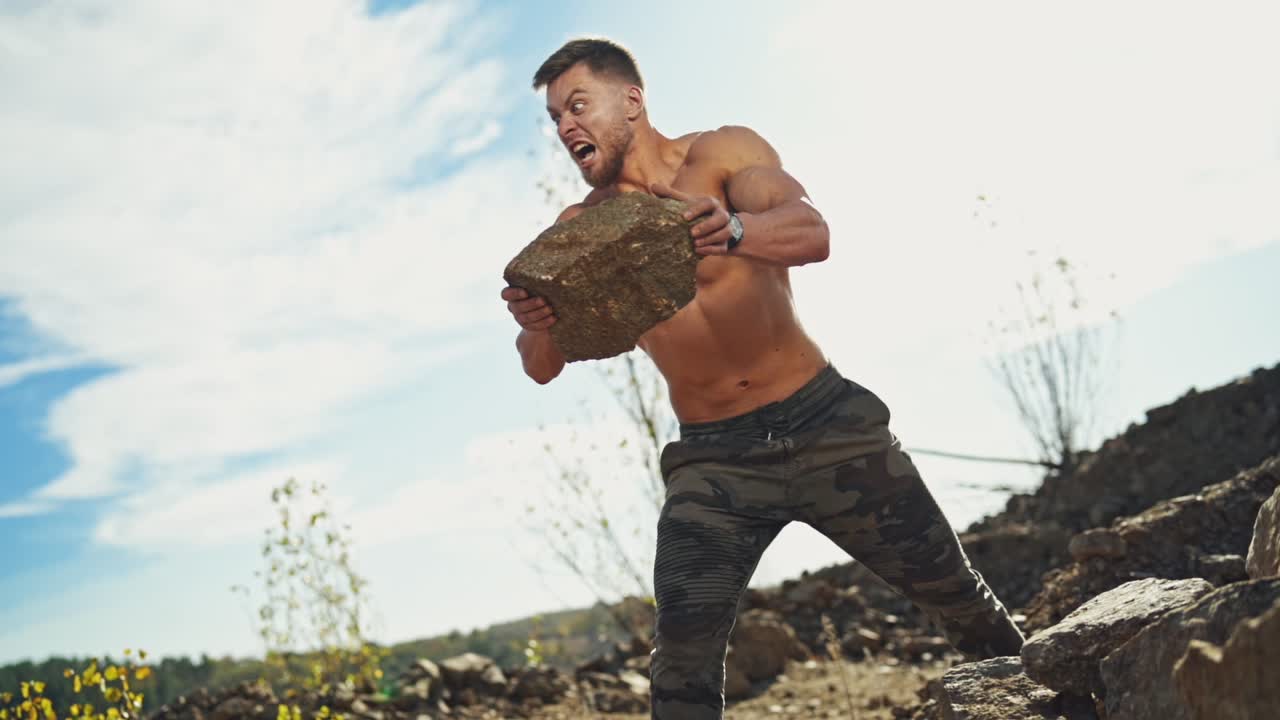Angry sportsman with muscular body throws stones in nature. Shirtless bodybuilder exercises with heavy stones outdoors. Slow motion.