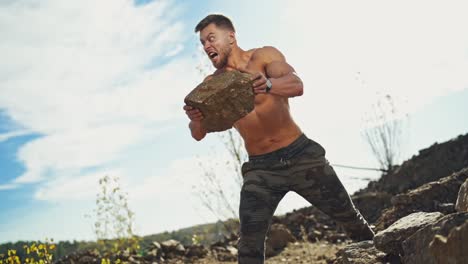 angry sportsman with muscular body throws stones in nature. shirtless bodybuilder exercises with heavy stones outdoors. slow motion.