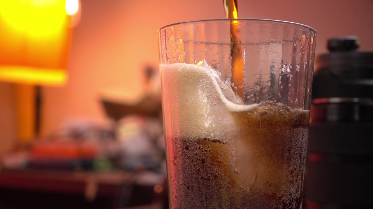 Soda versata in un bicchiere pieno di ghiaccio, che crea una schiuma in superficie e cattura bollicine in uno sfondo caldo e sfocato.