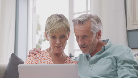 senior retired couple sitting on sofa at home shopping or booking holiday on laptop