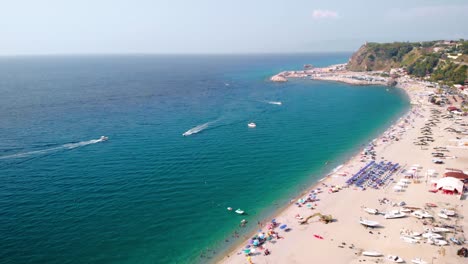 aerial, drone shot over the palmi beach, full of people, towards boats on the sea, sunny day, calabria, italy