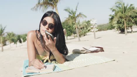 Fröhliche-Hispanische-Frau-Mit-Sonnenbrille,-Die-Am-Strand-Liegt-Und-Auf-Dem-Smartphone-Spricht,-Kopierraum,-Zeitlupe