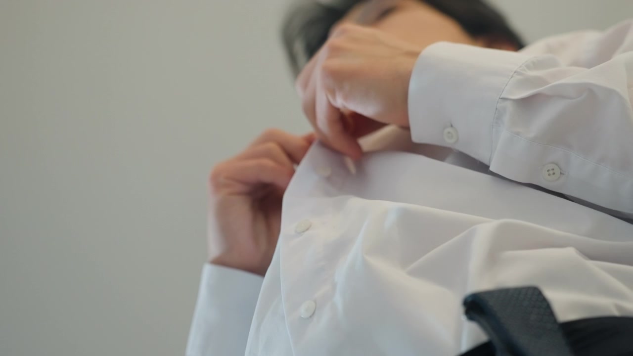 Uomo ansioso che abbottona la camicia bianca con mani tremanti, primo piano dal basso mostra la regolazione del colletto e del polsino, profondità di campo ridotta, postura rigida, si prepara come candidato per un colloquio fuori dalla hall dell'ufficio, pausa affannosa.