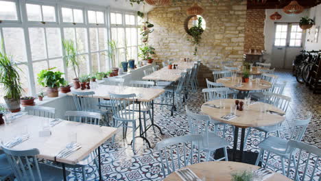 Toma-Manual-De-Mesas-Y-Sillas-Vacías-Junto-A-Una-Gran-Ventana-En-Un-Restaurante-Con-Plantas-De-Interior-En-El-Alféizar-De-La-Ventana,-Durante-El-Día