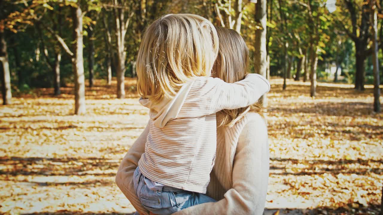 giovane madre bionda sorridente, tenendo in braccio la bambina e girando con lei durante la loro passeggiata nel parco d'autunno