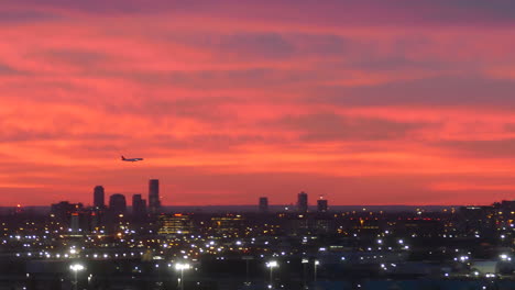 silhouette of plane landing above dusk cityscape with glowing lights and red sunset sky