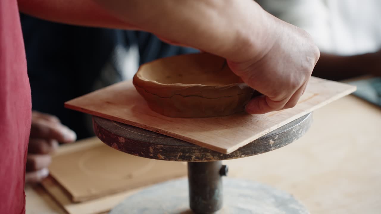 A close-up of a person's hands crafting a small, rustic bowl using a traditional hand-building pottery technique in a creative workshop or art class