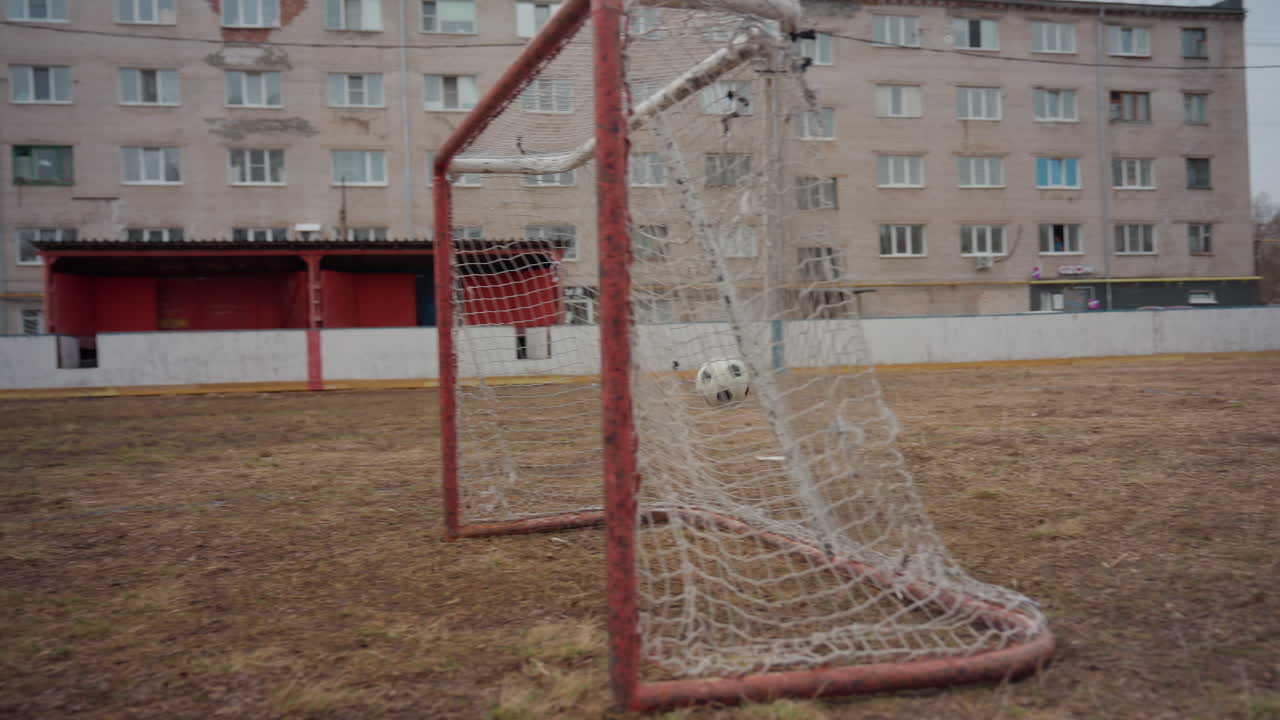 urban field showing white man goalkeeper practicing shots near rusted goal, empty apartment blocks framed by overcast sky, dry brown grass, solitary routine, nostalgic quiet atmosphere